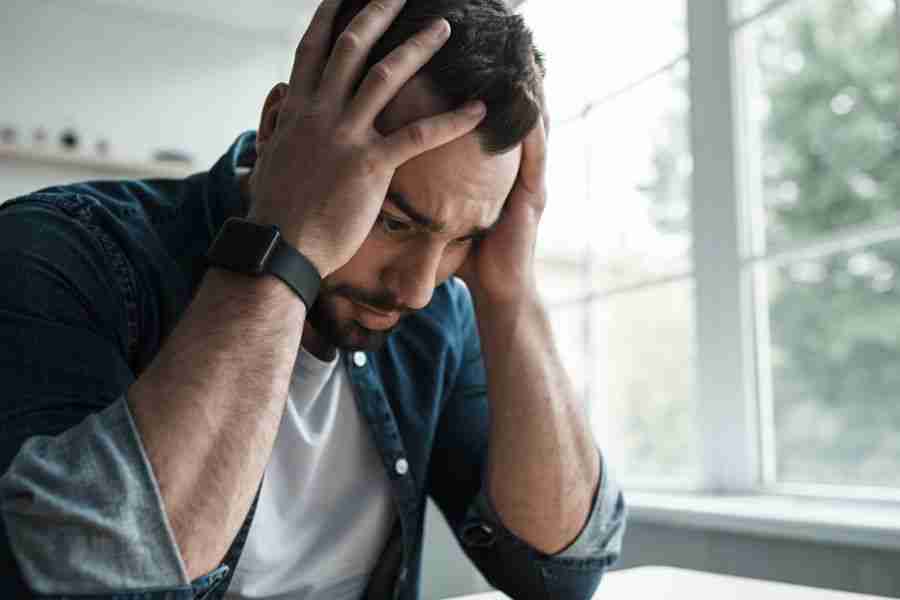 A man in a denim shirt and white t-shirt sits with his elbows on a table, holding his head in his hands. He appears distressed, perhaps wondering, Do I have anxiety? as natural light streams in through the nearby window.