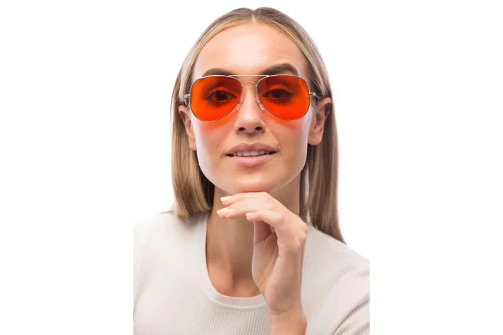 Holiday gifts for your favourite ADHD-brains 21 A woman with straight blonde hair, wearing a white top and orange-tinted aviator sunglasses, looks at the camera with a slight smile—her hand resting under her chin as if contemplating ADHD holiday gifts, set against a white background.
