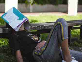 ADHD Essentials: 8 Therapist-Guided Tips for Thriving with Your ADHD Brain 3 A person lounging on a bench with a book covering their face, perhaps taking an ADHD essentials break. Theyre dressed in a black shirt with jeans that sport yellow and green stripes. The backdrop features a stretch of grass and part of a building, adding to the serene scene.