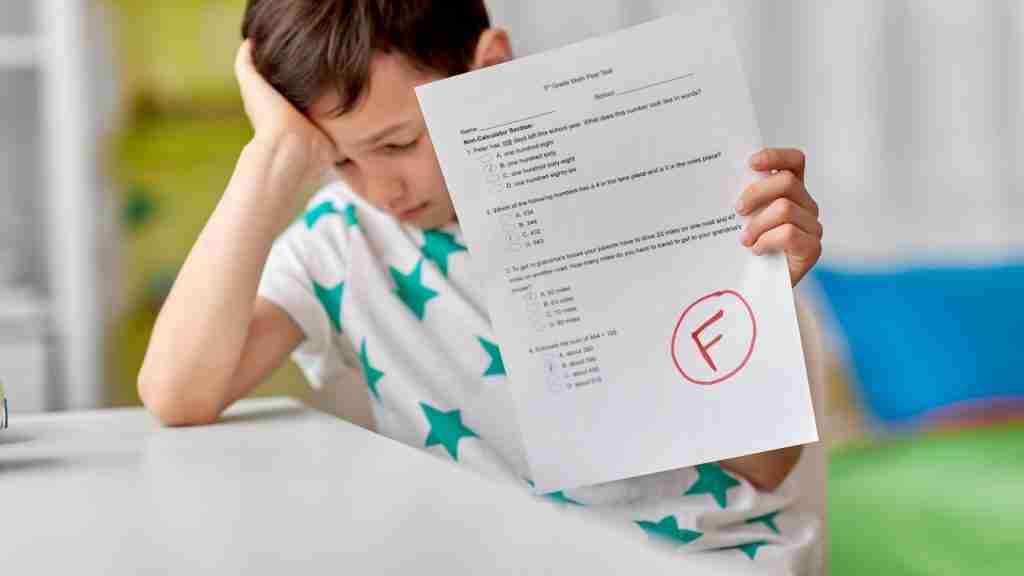 A young boy sits at a table, looking disappointed, holding a test paper with a large red F on it. Wearing a white shirt with green stars, he rests his other hand on his head, pondering what feels like The Reckoning, possibly facing late-diagnosed ADHD.