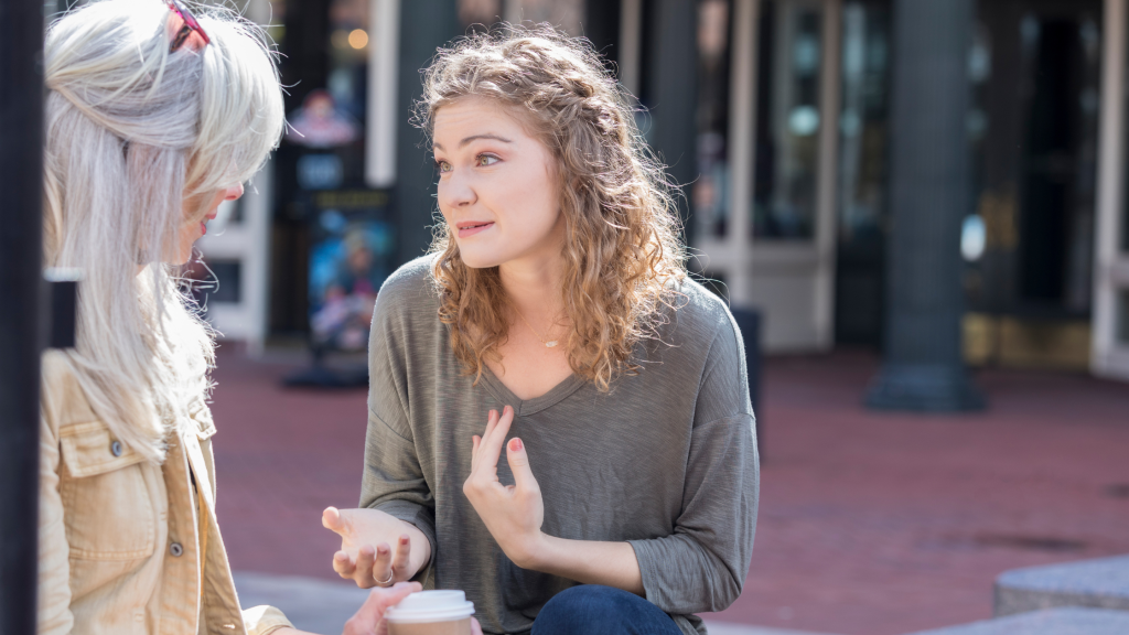Two women sit and converse outdoors. The woman on the right, with curly hair, gestures expressively while holding a coffee cup. Her companion, with long gray hair, listens attentively as they discuss parenting strategies for neurodivergent kids. Both appear deeply engaged in conversation.