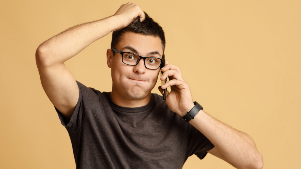 A person wearing glasses and a gray shirt holds a phone to their ear with one hand while scratching their head with the other. Set against a plain, warm beige background, they appear contemplative as if navigating family dynamics and neurodiversity.