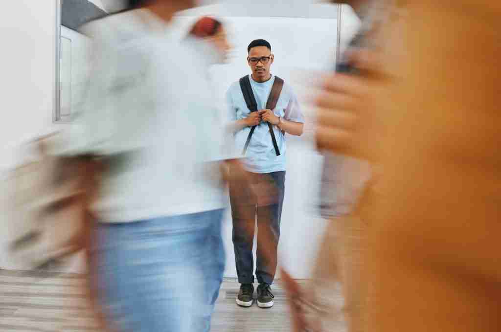 A person stands still against a wall, wearing glasses and a backpack. They appear contemplative as people in motion blur pass by, just like the calm assurance of Julia DAddurno RP offering therapist-approved AI to support your executive functioning amidst lifes chaos.