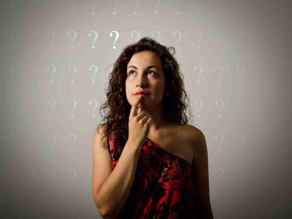 A woman with curly hair in a red and black top touches her chin, gazing upwards thoughtfully. The gray wall behind her is filled with question marks, symbolizing the curiosity and exploration often faced by ADHD families on their journey toward understanding and acceptance.