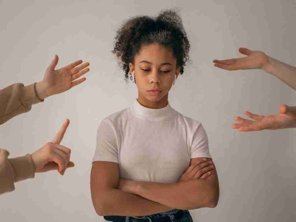 A young girl with curly hair stands with arms crossed, looking down. Multiple hands from outside the frame gesture toward her, capturing the pressure she feels. This moment reflects conversations families need to have about ADHD acceptance, fostering understanding and support.