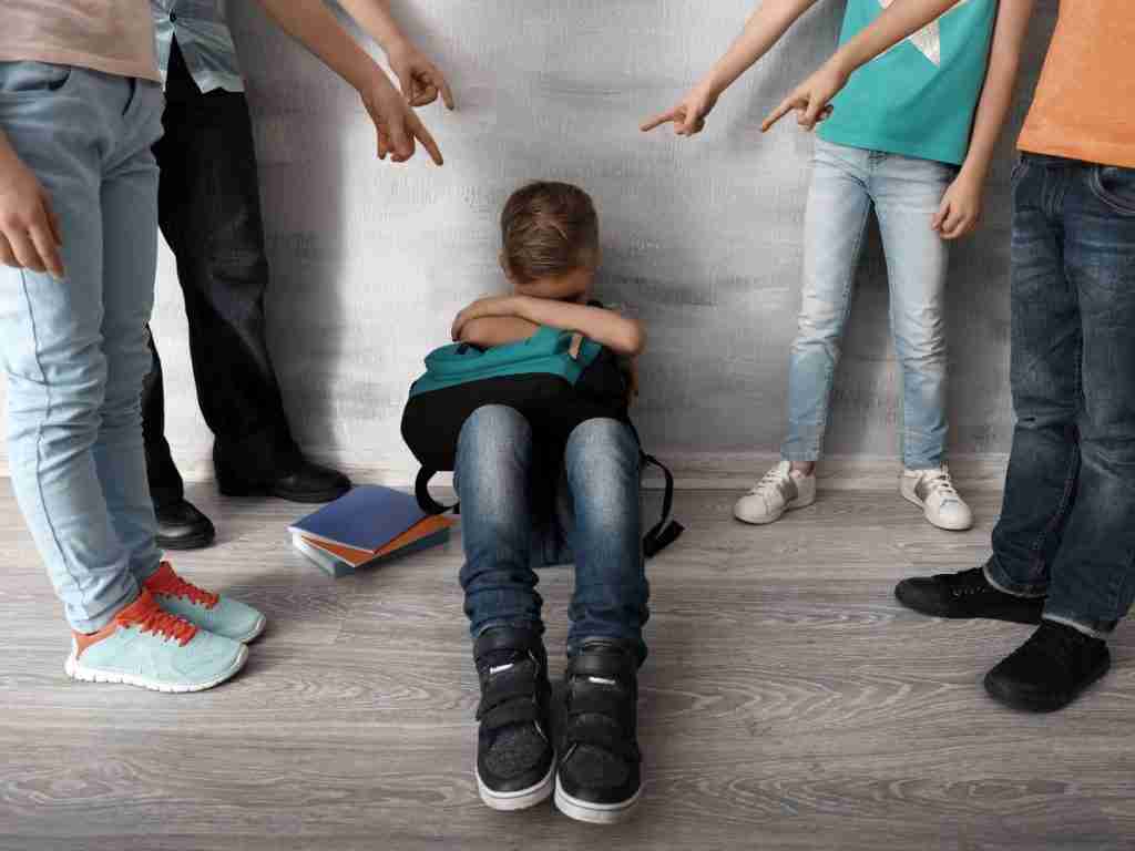 A child, possibly struggling with autism or ADHD, sits on the floor against a wall, hugging their knees as peers point at them. Distressed, they are surrounded by scattered books. This scene underscores the importance of family support and understanding neurodiversity in parenting.