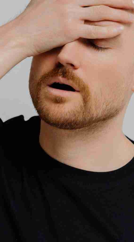 A person with a beard and mustache is wearing a black shirt. With one hand on their forehead and eyes closed, they seem deep in thought or frustrated. Their expression hints at the challenges of navigating ADHD and ableism, seeking understanding amidst a plain, light-colored backdrop.