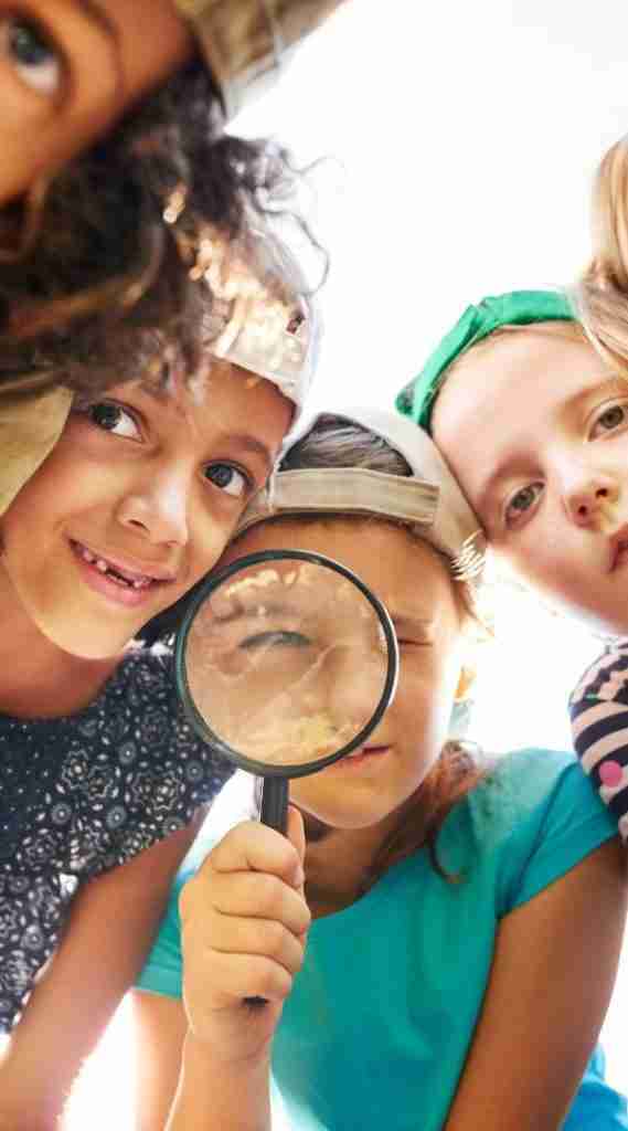Children in a close group, one holding a magnifying glass in front of her face, distorting her features. Theyre outdoors, embracing neurodiversity and parenting with playful curiosity. Casual clothing and caps complement their joyful expressions as the bright sunlight shines on their cheerful scene.
