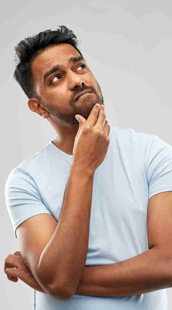A man in a light blue t-shirt stands against a plain gray background, looking up contemplatively with his hand on his chin. His thoughtful expression might reflect on the challenges of ADHD and ableism in everyday life.
