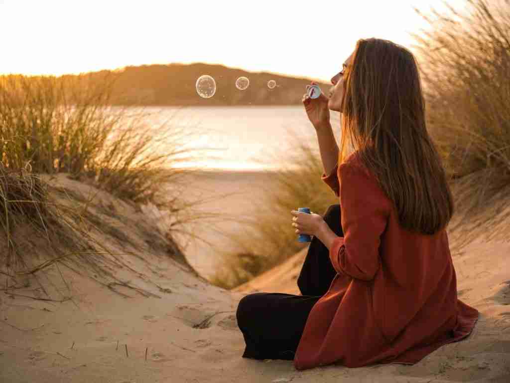 A woman practices mindfulness as she sits on sand among tall grass at sunset, blowing soap bubbles, with a calm body of water and hills in the background.