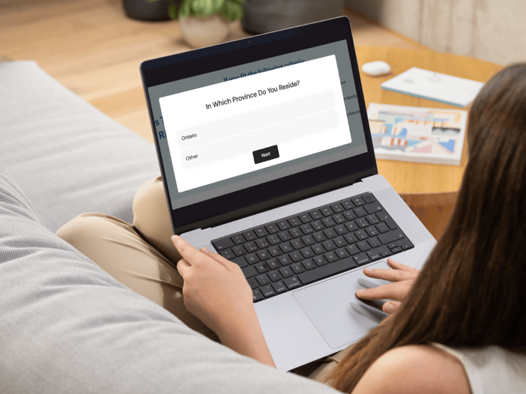 A person sits on a couch using a laptop displaying a form that asks, In which province do you reside?—with options for Ontario and Other—while searching for private psychiatry Ontario services when the wait feels too long.