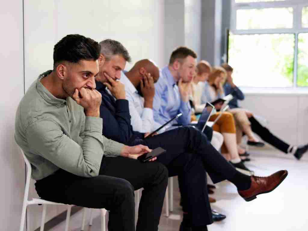 A row of people sitting on chairs in a waiting room, all looking anxious or deep in thought as the wait feels too long; some hold phones or papers, while daylight streams through a window in the background.