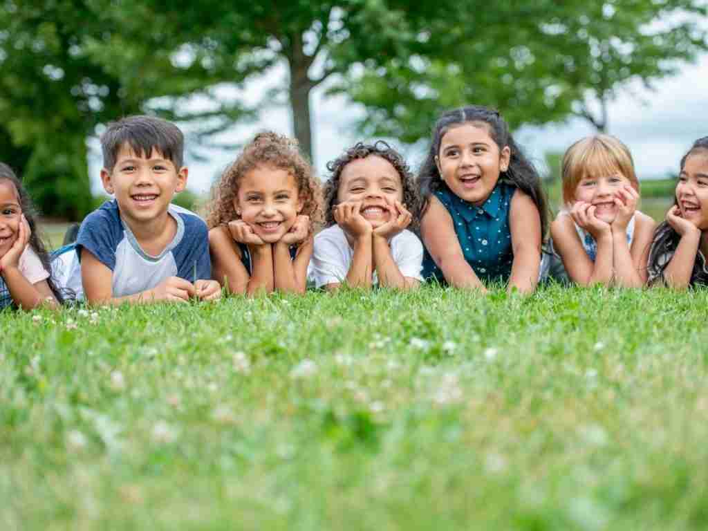 Seven young children lie on their stomachs in the grass, smiling and resting their faces in their hands, showing how to feel less frazzled and enjoy summer relaxation with trees and greenery in the background on a bright day.