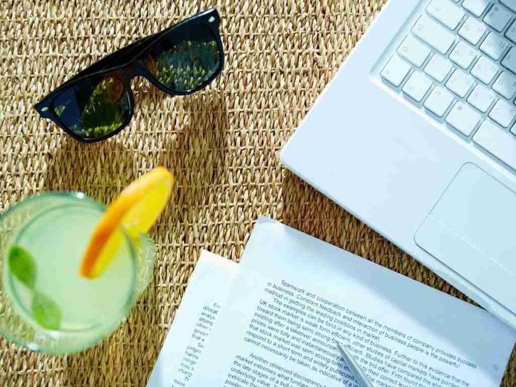 A glass of lemonade with mint and an orange slice, a pair of sunglasses, papers, and a laptop rest on a woven surface—capturing the essence of summer and stress management in a relaxed work or study setting.