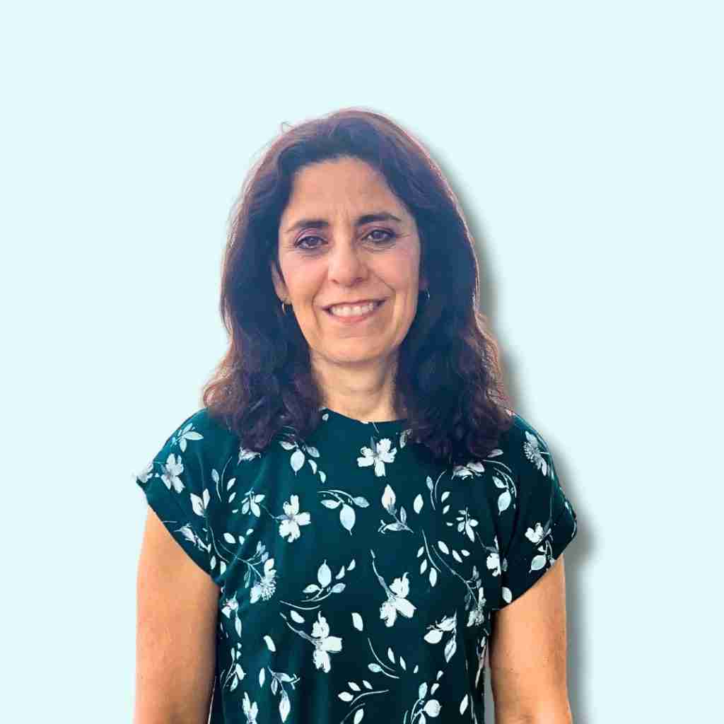 A woman with wavy brown hair, wearing a short-sleeved, dark green floral top, stands smiling in front of a plain light blue background, representing the Leadership team at Dig a Little Deeper Ontario.