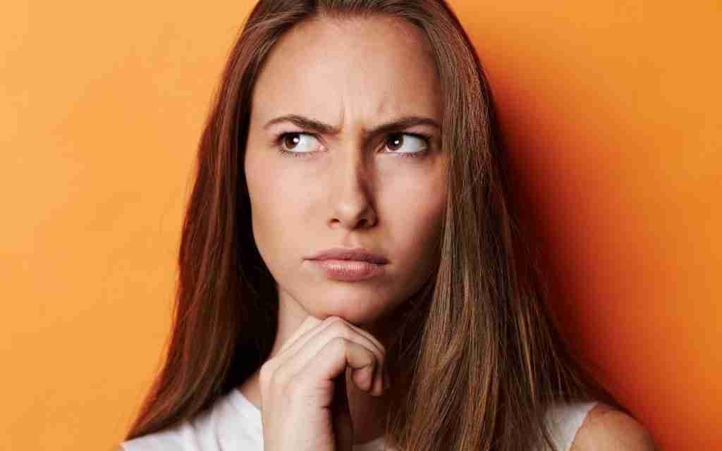 A woman with long brown hair looks up and to the side with a furrowed brow, appearing thoughtful or confused—perhaps pondering how to start practicing self-compassion—while resting her chin on her hand. The background is orange.