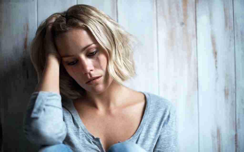 A woman with short blonde hair sits against a wooden wall, resting her head on her hand and looking down with a sad expression, perhaps reflecting on self-compassion as she navigates the challenges of ADHD, dressed in a gray top.