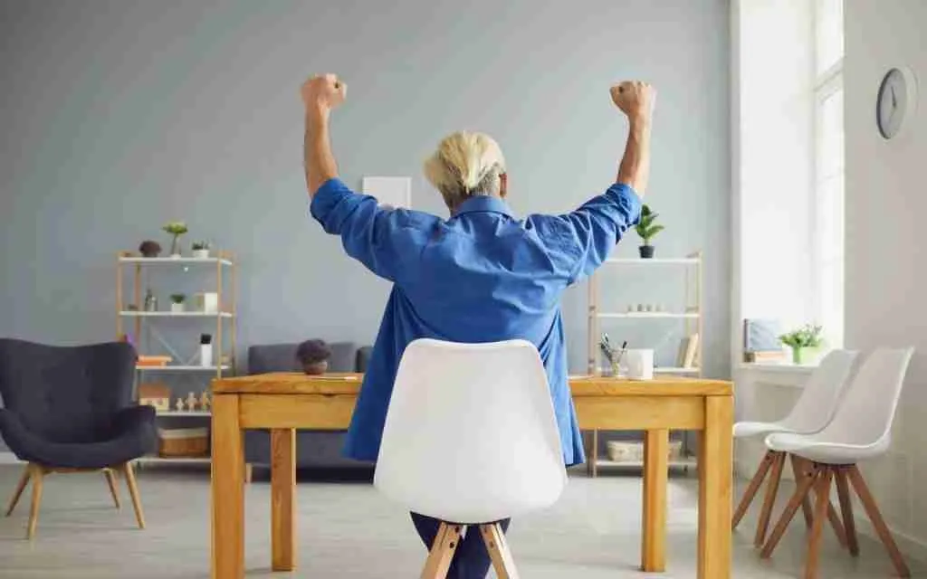 A person with blond hair sits at a desk facing away, raising both arms in celebration—an uplifting moment of self-compassion. The room features shelves, plants, and a window letting in natural light.