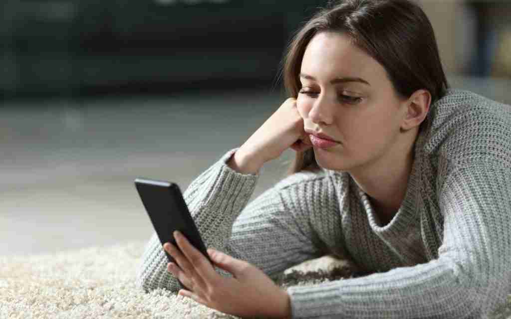 A young woman lies on a light-colored carpet, propped on one elbow, looking at her smartphone with a bored expression—a scene reflecting underarousal ADHD and common task initiation problems as she struggles to find the motivation to get started.
