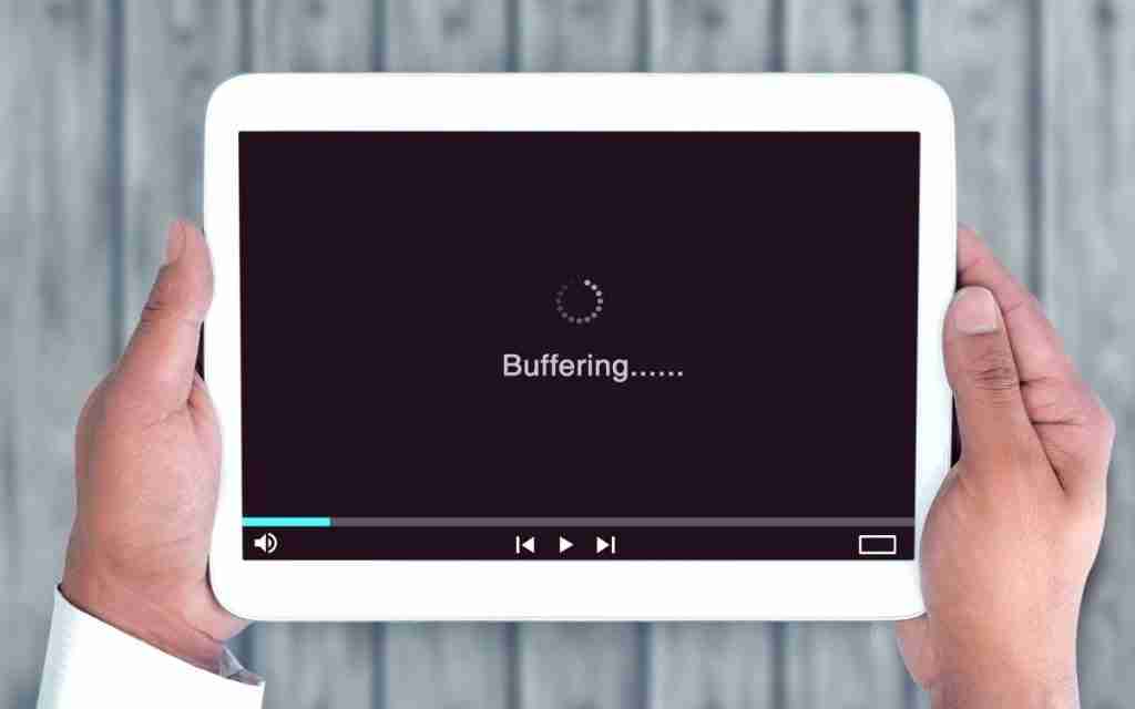 A person holds a white tablet displaying a video player screen with the word Buffering...... and a loading icon, symbolizing ADHD and low activation state. The background features a wooden surface.