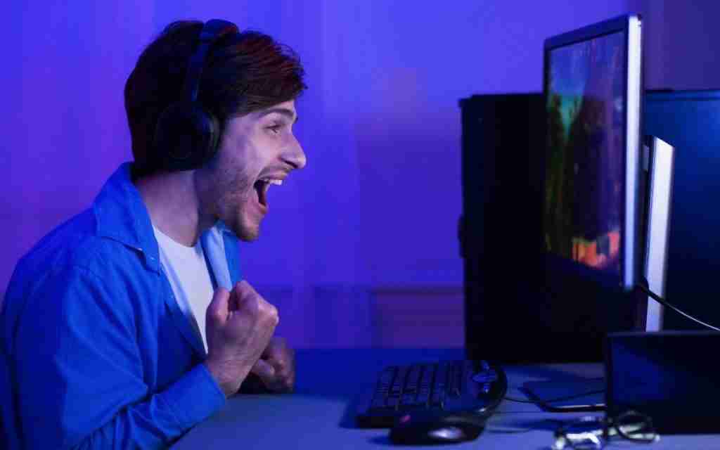 A young man wearing headphones and a blue shirt excitedly cheers while looking at a computer monitor, overcoming ADHD task initiation problems in a room lit with purple light, with a keyboard and mouse in front of him.