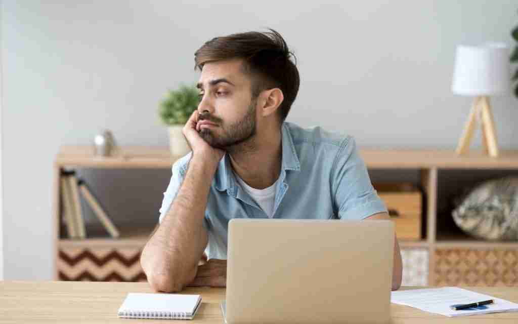 A man sits at a desk with a laptop, notebook, and pen, resting his chin on his hand and staring off to the side—his expression reflecting inattentive ADHD motivation or an ADHD and low activation state in a home office setting.