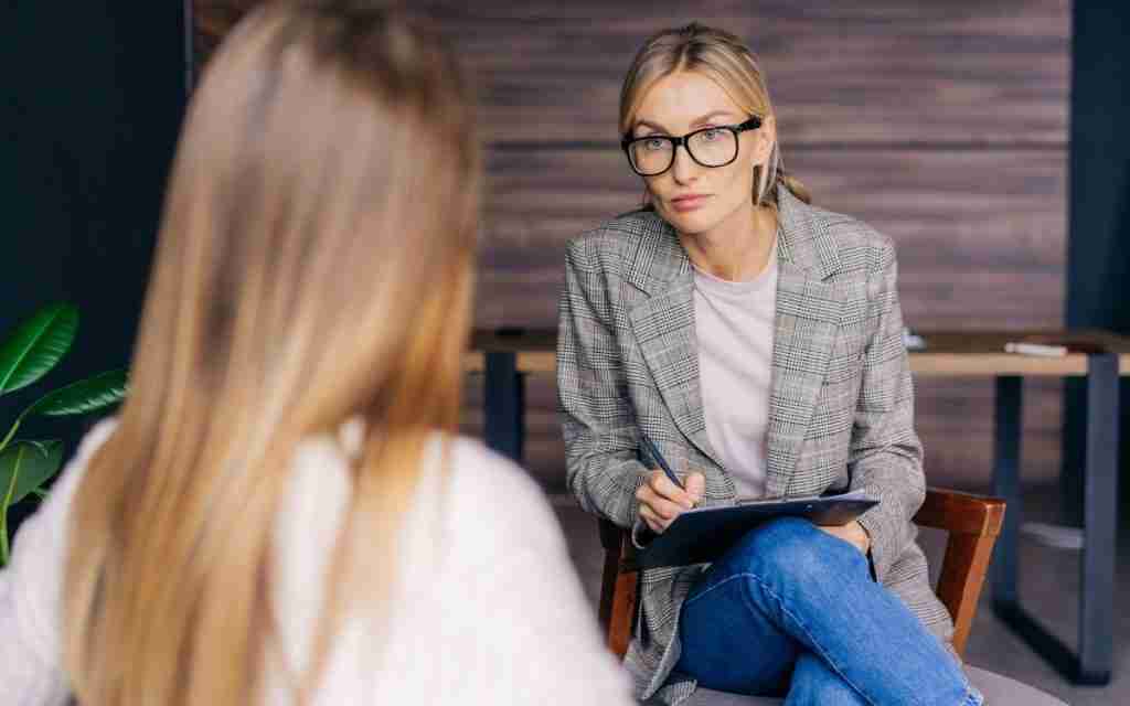A woman wearing glasses and a plaid blazer, possibly a DIRECTOR INTEGRATED MENTAL HEALTH or PMHN, sits with a clipboard and pen, attentively listening to another woman with long hair during an ADHD diagnosis Ontario office consultation.