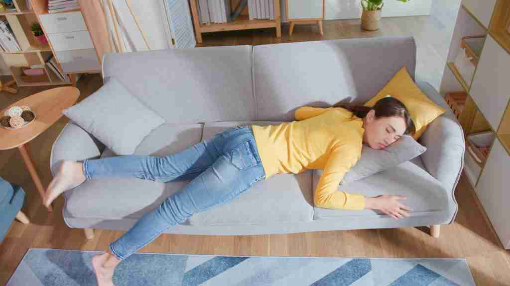 A young woman in a yellow sweater and jeans is lying face down on a gray sofa, appearing tired or asleep from midlife fatigue, with her head on a pillow and one leg dangling off the couch in a cozy living room.