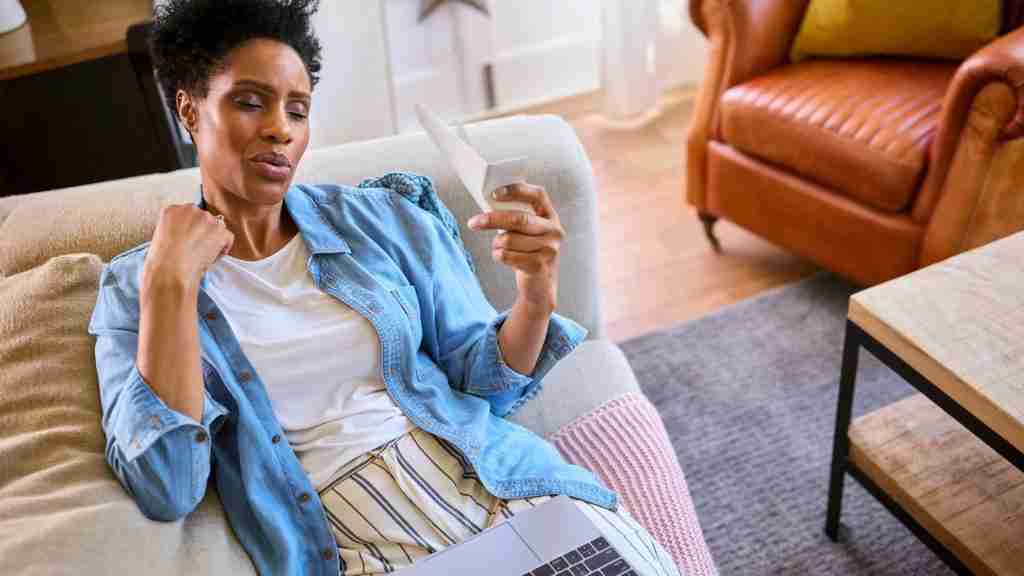 A midlife woman sits on a couch, looking at a piece of paper with concern, her laptop on her lap in a cozy living room—perhaps reflecting on perimenopause or ADHD symptoms.