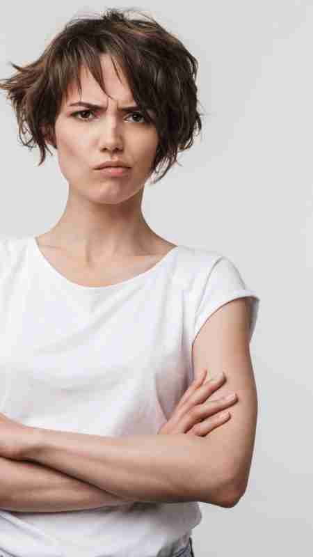 A young woman with short, tousled brown hair wearing a white t-shirt stands with her arms crossed, looking forward with a serious and slightly angry expression, reflecting the challenges of midlife.