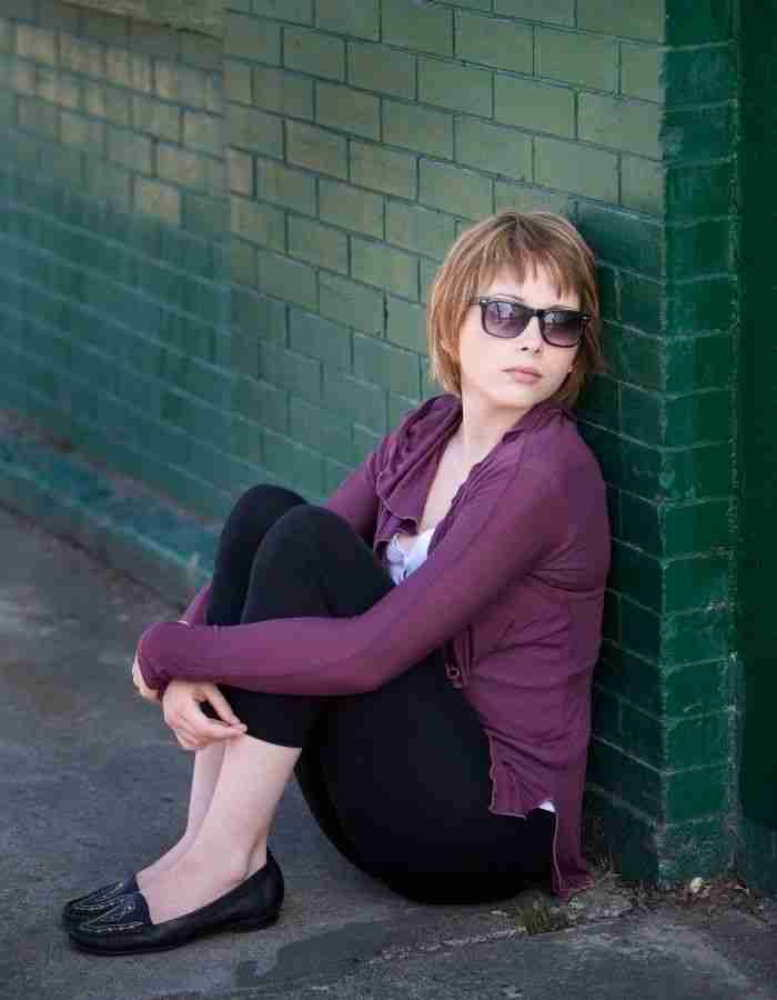 A person with short light hair, wearing sunglasses, a purple cardigan, black leggings, and black shoes, sits on the ground leaning against a green brick wall outdoors—reflecting the quiet perseverance often found in High Achievers ADHD and mental health journeys.