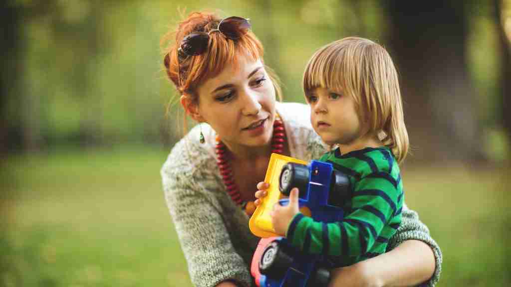 A woman with red hair and sunglasses on her head crouches next to a young child holding a toy truck, enjoying time together outdoors in a green, park-like setting that promotes mental health.