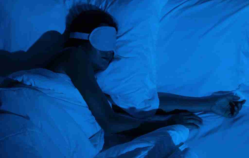 A person lies in bed under white blankets, wearing a sleep mask and resting on a pillow, bathed in blue-tinted light, appearing to quiet your mind and find rest after sleepless nights.