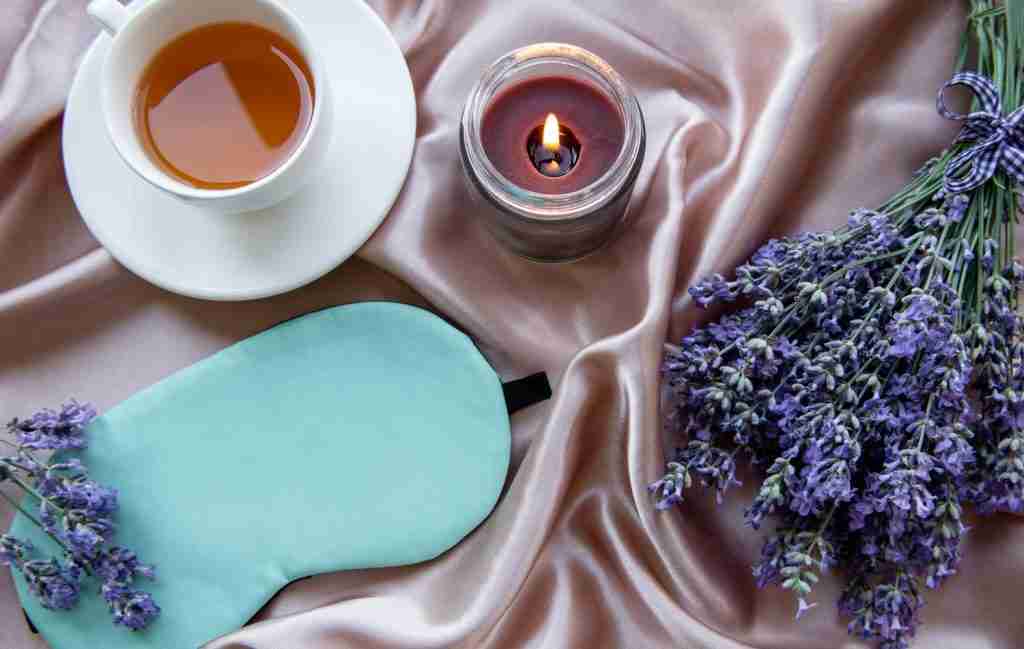 A cup of tea, a lit candle, a green sleep mask, and fresh lavender rest on pink satin fabric—perfect to quiet your mind and bring calm after sleepless nights.