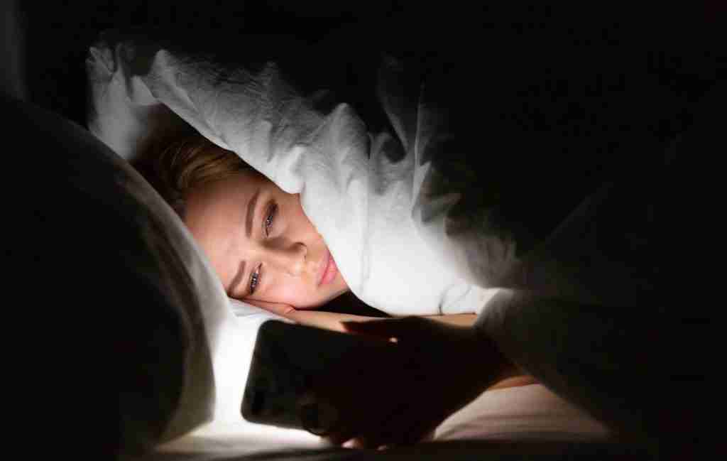 A person lies in bed under a blanket at night, looking at a smartphone, the screen illuminating their face—another of those sleepless nights where it's hard to quiet your mind.