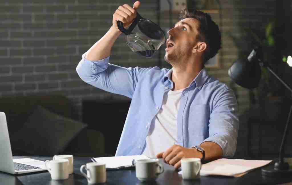 A man with sleepless nights sits at a desk piled with empty coffee cups, tilting a coffee pot above his mouth. He looks tired and overwhelmed, suggesting he's struggling to quiet his mind after a long, stressful work session.