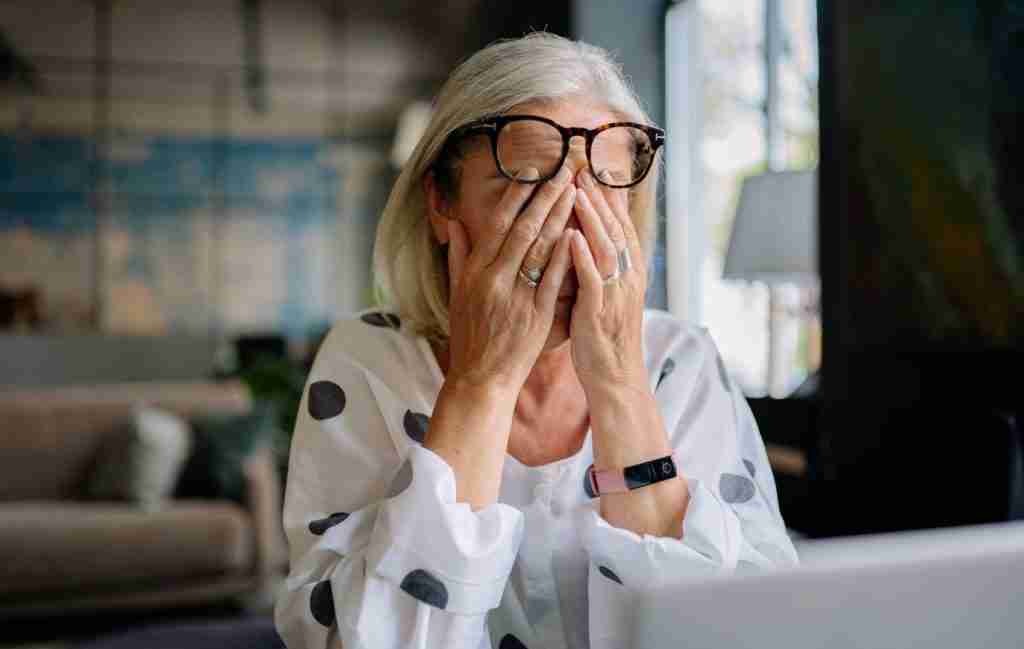 An older woman with gray hair and glasses sits at a table with her hands covering her face, appearing stressed or tired. She’s wearing a white shirt with large polka dots, a fitness tracker, and showing the gap in awareness about ADHD in seniors.