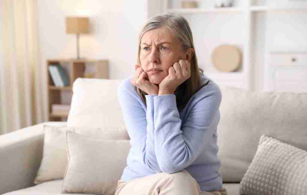 An older woman with gray hair sits on a beige sofa, resting her chin on her hands and looking thoughtfully into the distance. In this bright living room, she reflects on elderly mental health while wearing a light blue sweater.