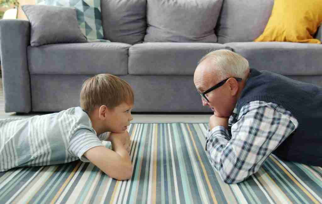 A young boy and an older man lie facing each other on a striped rug, smiling and talking about ADHD, their heads propped on their arms. A gray couch with cushions is in the background. The scene bridges the gap between generations.