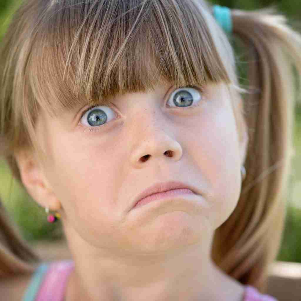 A young girl with blue eyes and light brown hair in pigtails looks surprised or confused, her wide eyes mirroring the gap in understanding often seen in ADHD. The background is blurred greenery.