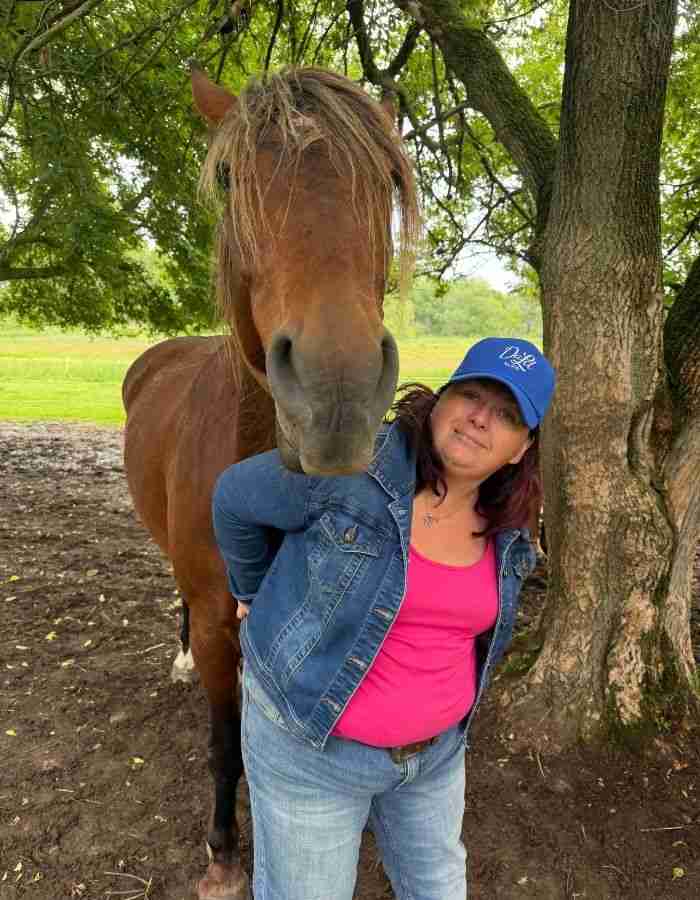 Finding the Right Kind of Horse-Assisted Support: What to Know Before You Book 3 A woman in a blue hat and denim jacket stands next to a brown horse under a tree, smiling at the camera. The pair enjoy the outdoors in a grassy, wooded area—an inviting scene that reflects the calm connection found in equine therapy.
