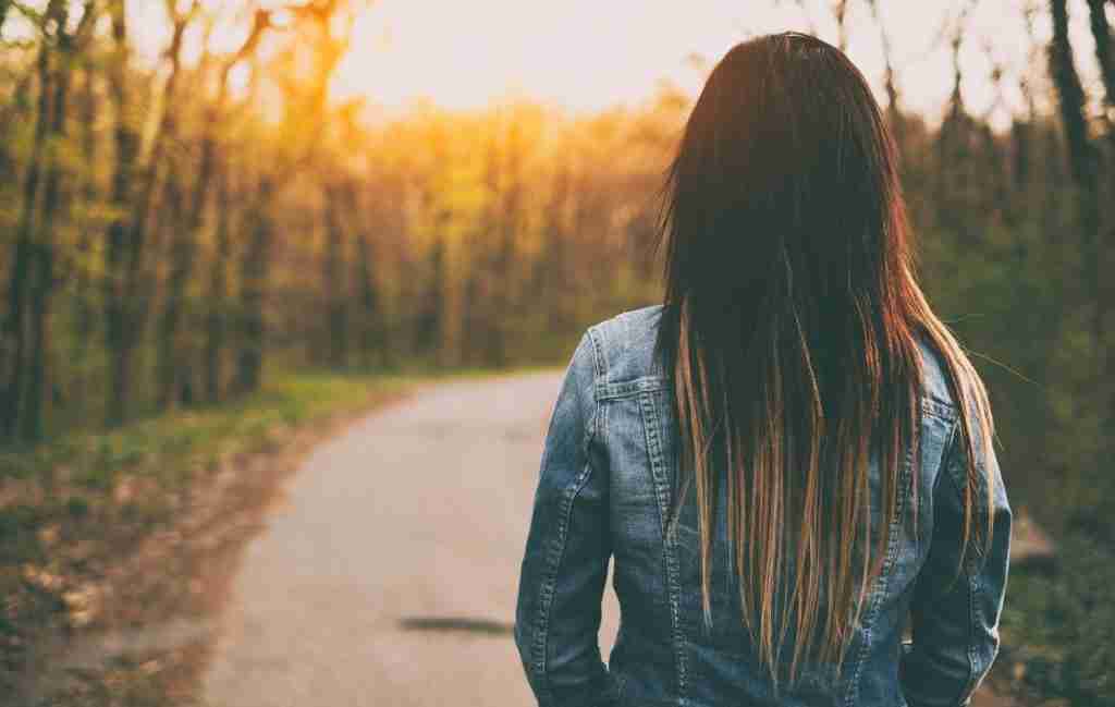 A person with long hair wearing a denim jacket stands on a path in a forest, facing away toward the setting sun, reflecting on personal growth and the values that guide their journey.
