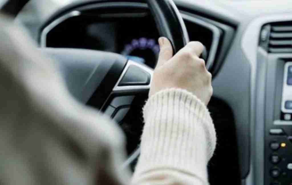 A person wearing a light-colored sweater grips a car steering wheel, embodying decision making as they navigate the road ahead, with part of the dashboard and infotainment system visible in the background.