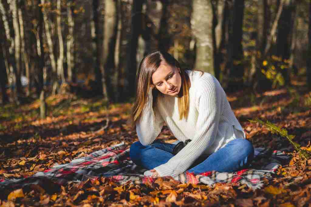 A woman in a white sweater and jeans sits on a plaid blanket in a forest, surrounded by autumn leaves and sunlight filtering through the trees—a tranquil setting perfect for quiet reflection on personal growth and values.