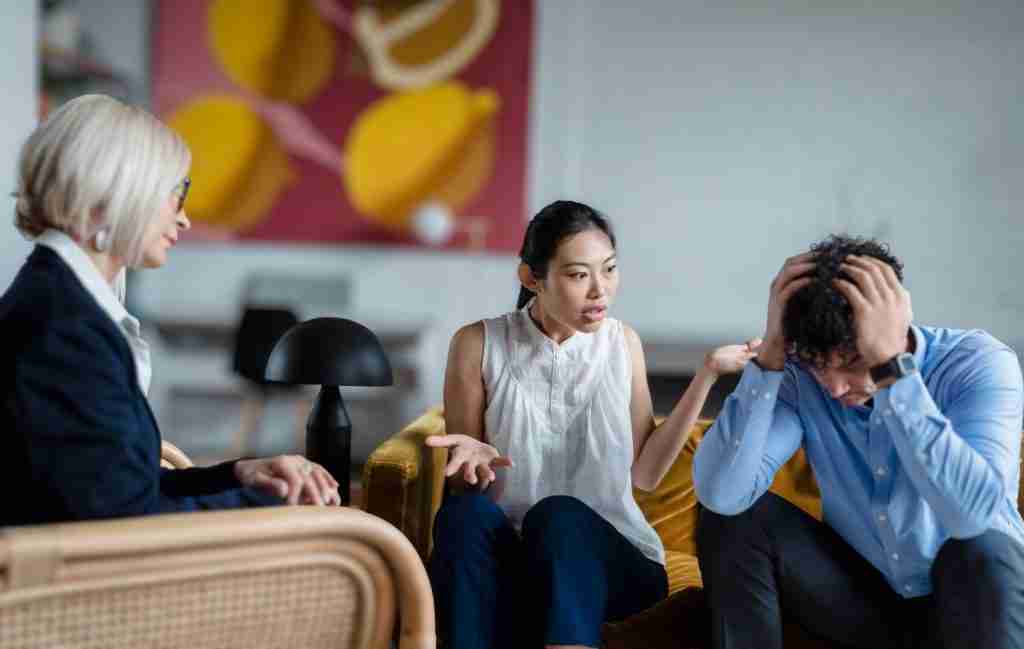 Three people sit indoors; a woman in white gestures emotionally while talking to a distressed man holding his head. Another woman listens attentively. The mood appears tense, suggesting a counseling or mediation session centered around ADHD communication problems and their impact on relationships.
