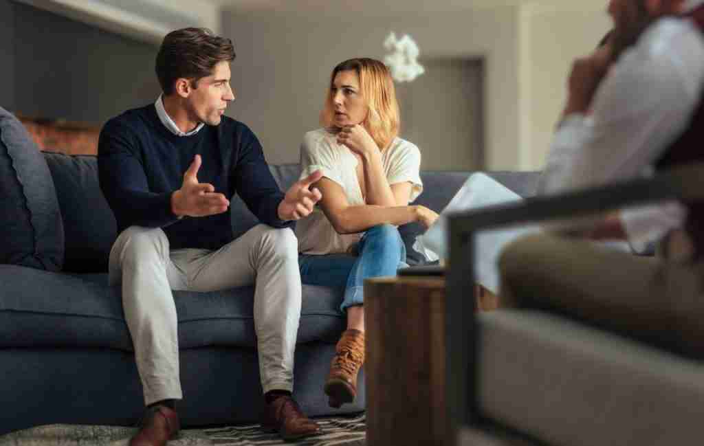 A man and woman sit on a couch having a serious conversation, while another person sits across from them, suggesting a counseling or therapy session in a comfortable living room setting. The discussion appears focused on addressing ADHD communication problems and how slow processing speed may impact their relationship dynamics.